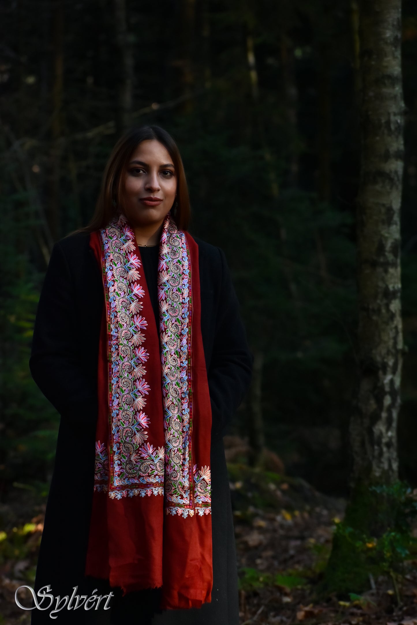 A female model in a woodland setting wearing a floral bordered red Rivan Cashmere stole from Sylvért's Rivanor collection
