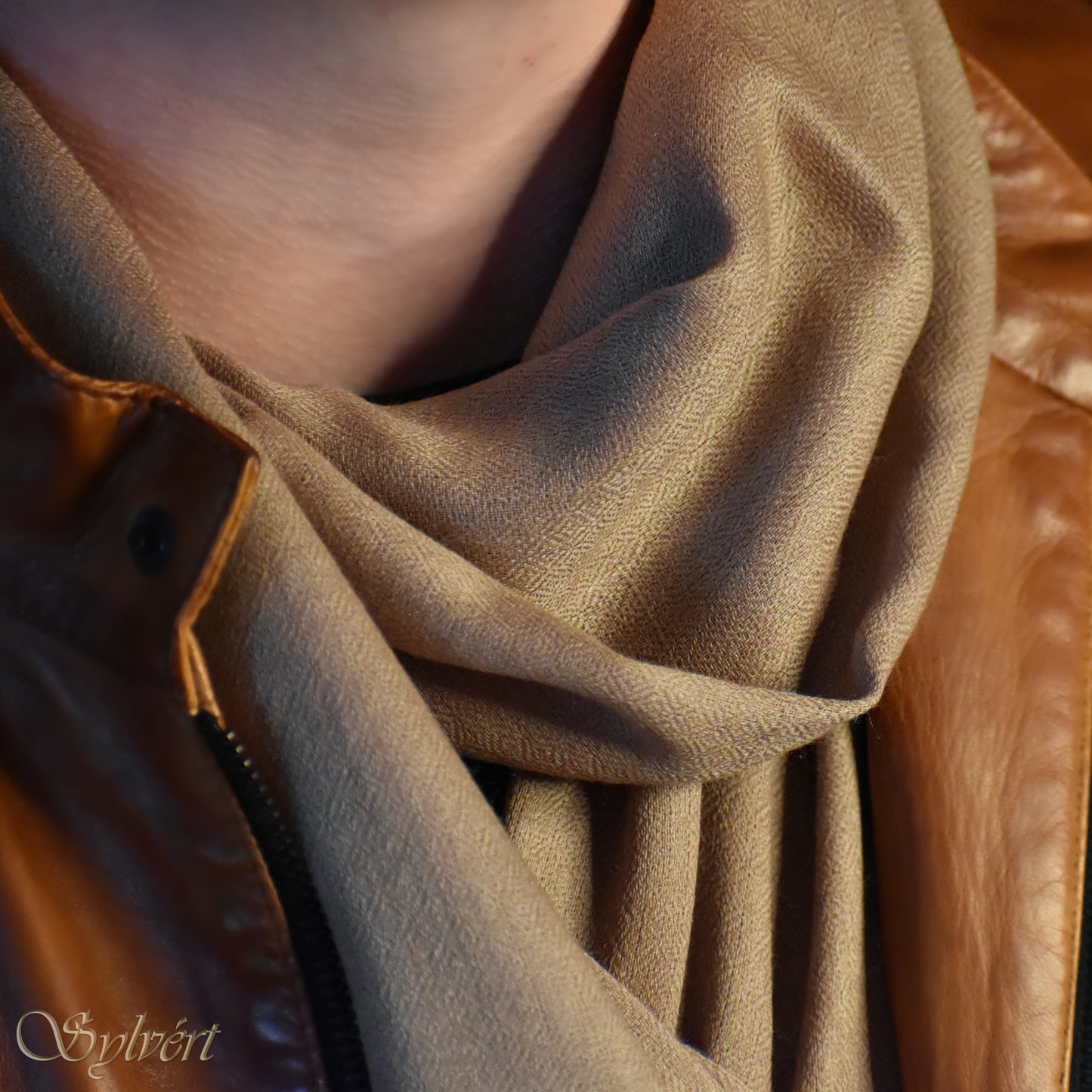 A styled shot of a natural blush cashmere Rivella Relor scarf from Sylvért's Rivanor collection worn under a brown leather jacket