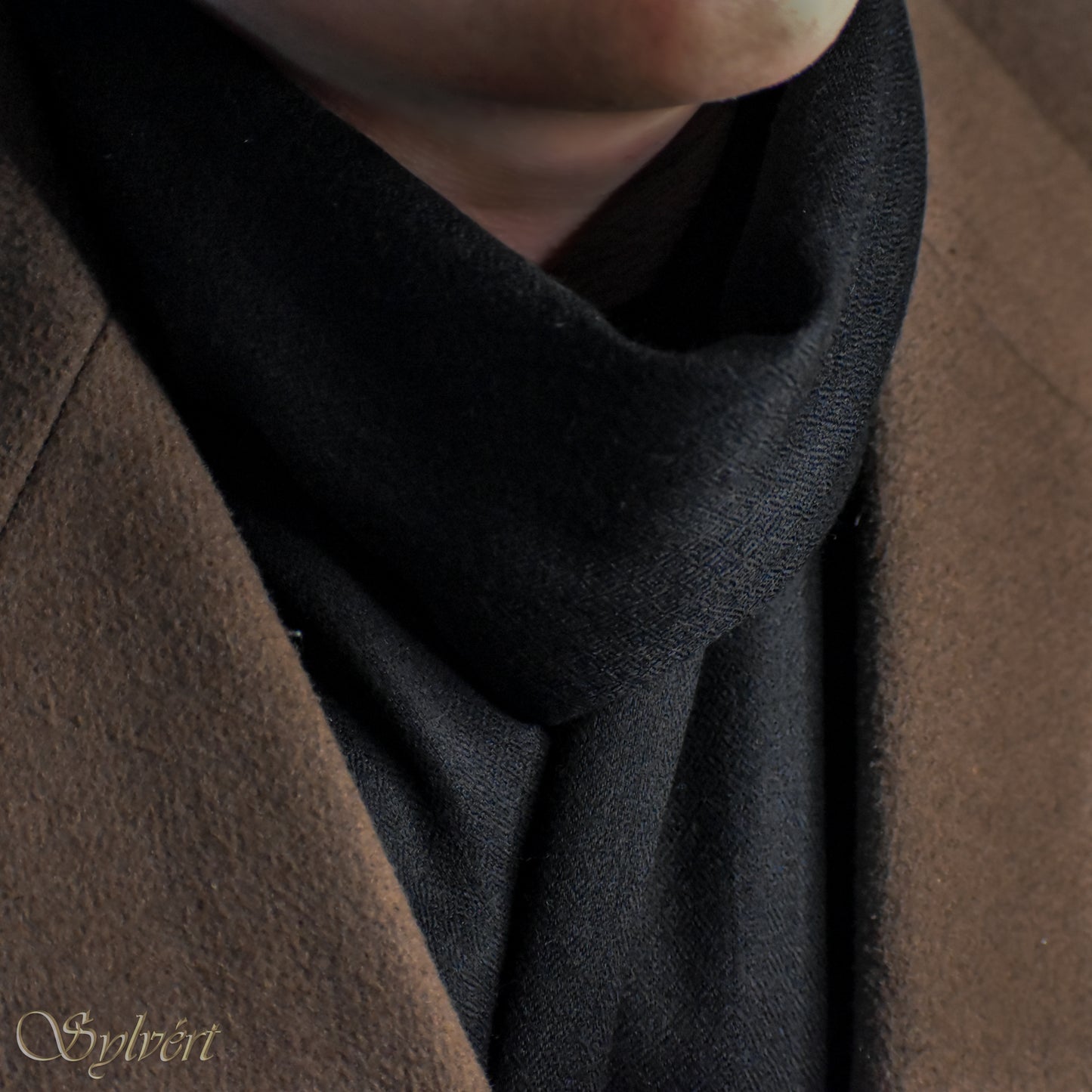 A styled shot of a black cashmere Rivella Velin scarf from Sylvért's Rivanor collection worn under a brown coat