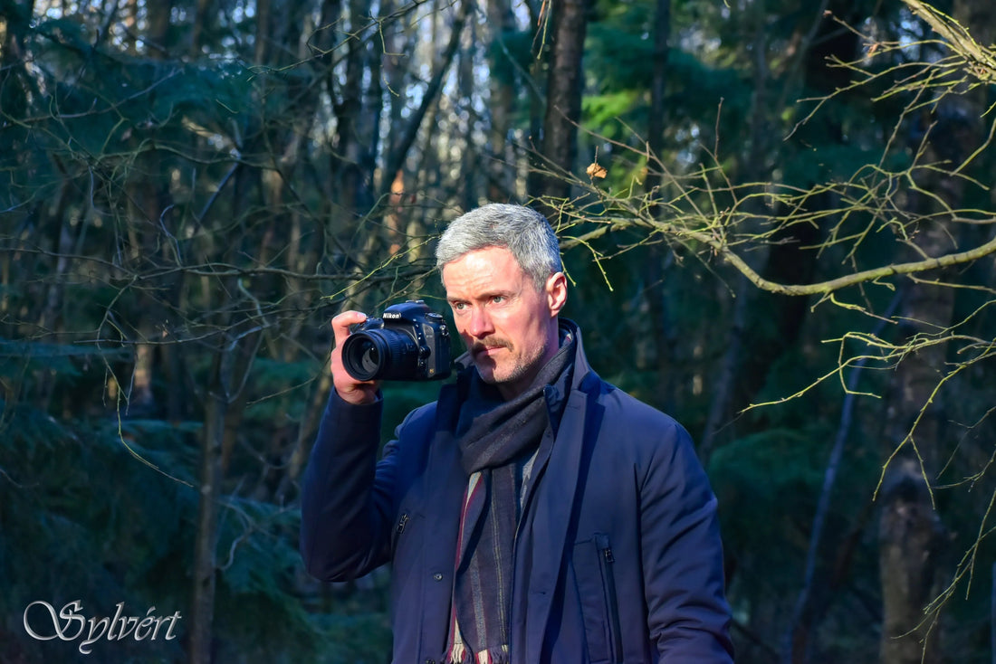 Middle-aged man with gray hair holding a Nikon camera in a sunlit forest wearing a dark coat and scarf