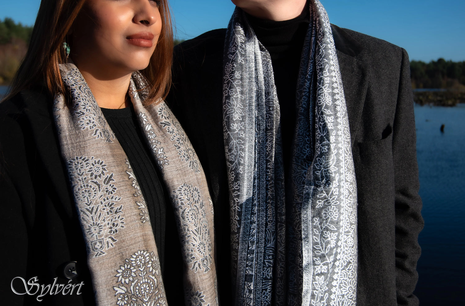 Two people wearing black coats and floral patterned scarves near a lake under clear blue sky