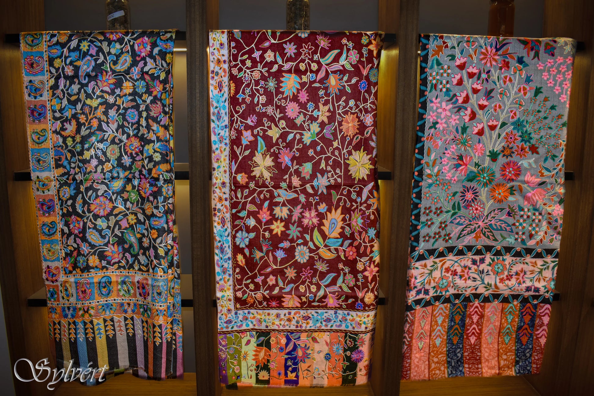 Three colorful floral embroidered shawls draped on wooden shelves with intricate patterns and stripes