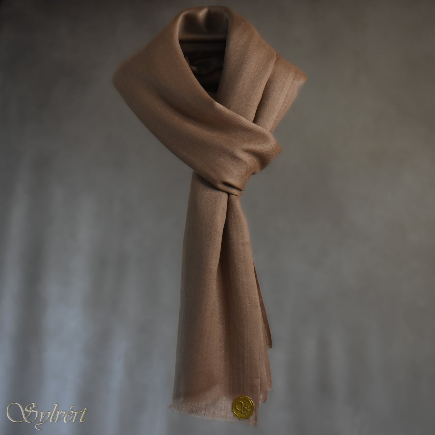 A natural "blush" cashmere Rivella Relor Scarf from Sylvért's Rivanor Collection. looped and threaded, suspended in the air in front of a beige background showing the Sylvért Stamp