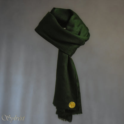 A green cashmere Rivella Tavon Scarf from Sylvért's Rivanor Collection, looped and threaded and showing the gold Sylvért stamp, suspended in the air in front of a beige background