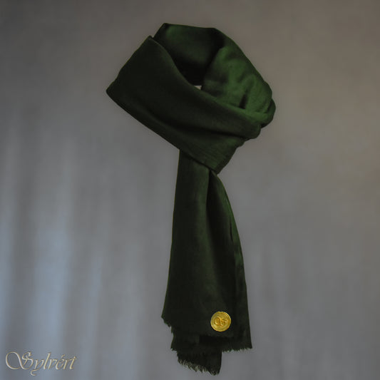 A green cashmere Rivella Tavon Scarf from Sylvért's Rivanor Collection, looped and threaded and showing the gold Sylvért stamp, suspended in the air in front of a beige background