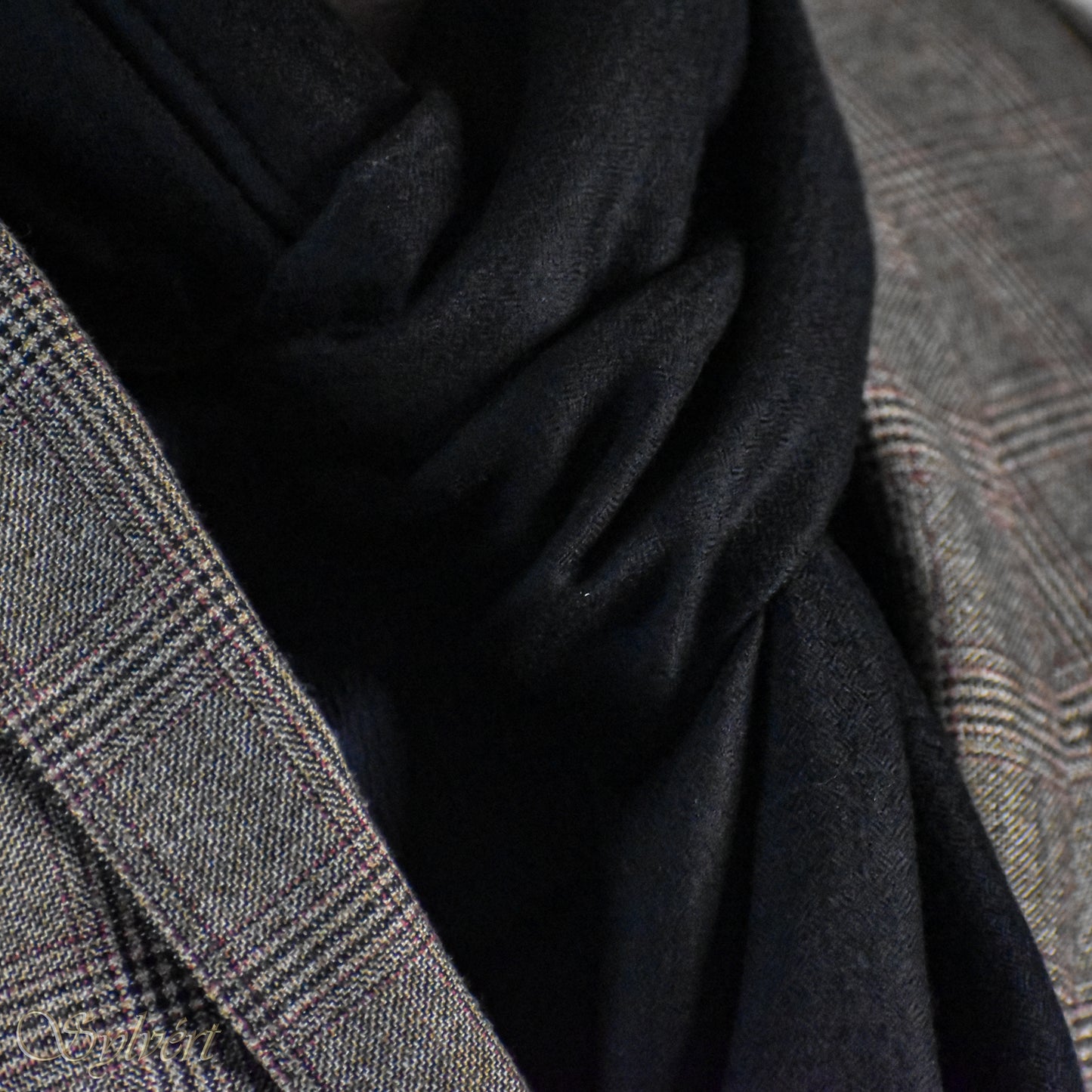 A styled showcase of a black Corvella Noctra from Sylvért's Coronis Collection, worn snugly under a grey plaid blazer
