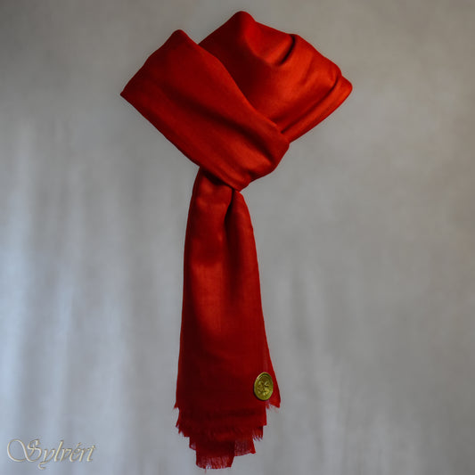 A red cashmere Rivella Sirel Scarf from Sylvért's Rivanor Collection, looped and threaded showing the gold Sylvért stamp, suspended in the air in front to of a beige background