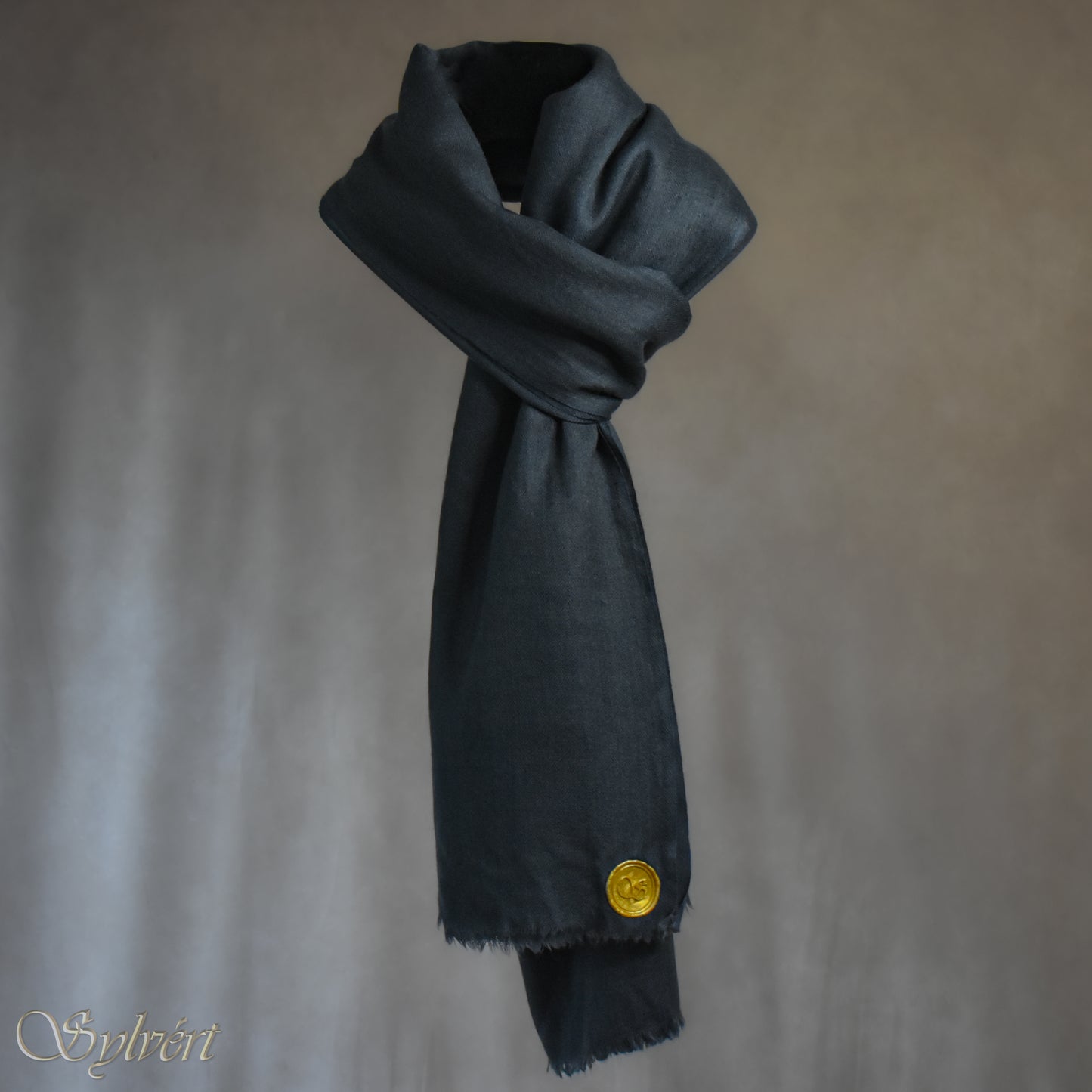 A blue-grey slate cashmere Rivella Veyra Scarf from Sylvért's Rivanor Collection, looped and threaded showing the gold Sylvért stamp, suspended in the air in front to of a beige background