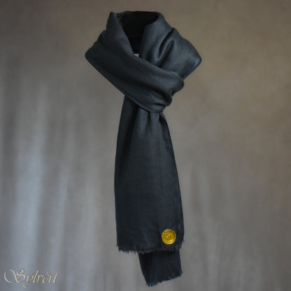 A blue-grey slate cashmere Rivella Veyra Scarf from Sylvért's Rivanor Collection, looped and threaded showing the gold Sylvért stamp, suspended in the air in front to of a beige background