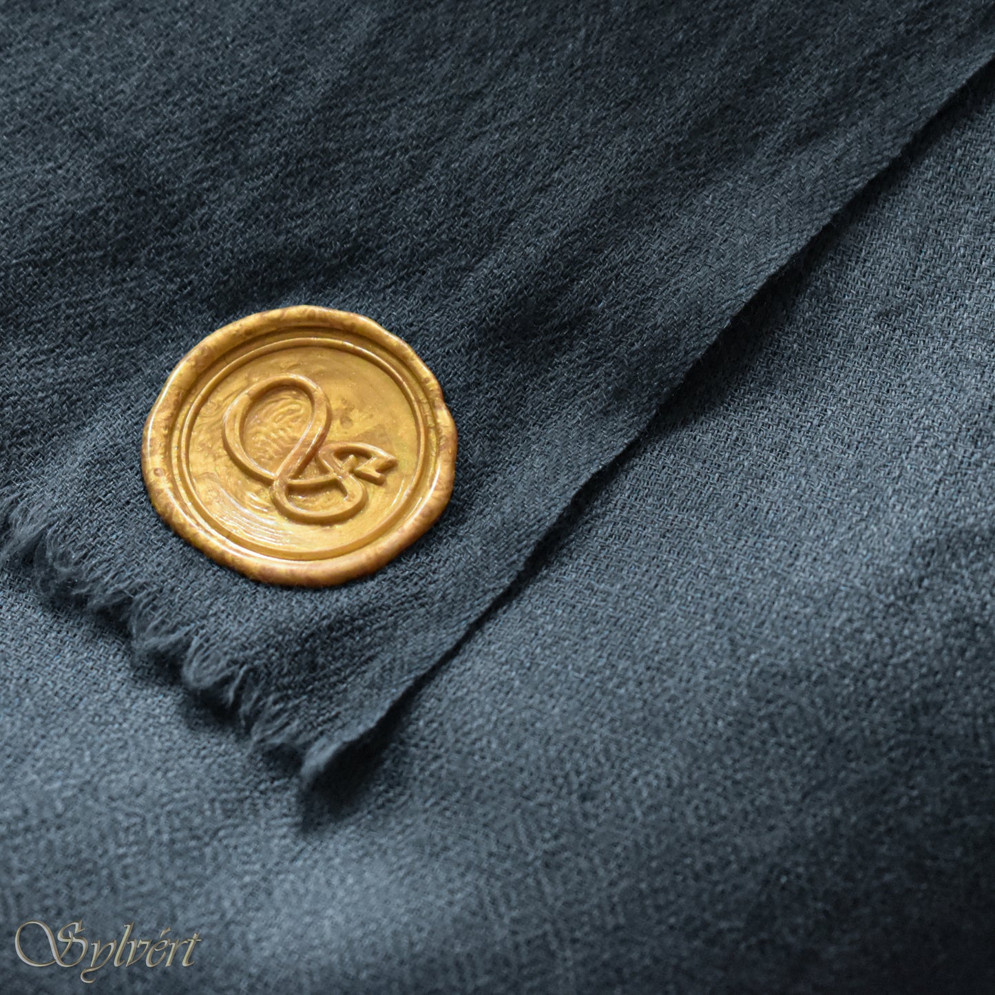 A blue-grey slate cashmere Rivella Veyra Scarf from Sylvért's Rivanor Collection, folded to show the diamond weave texture and the gold Sylvért stamp