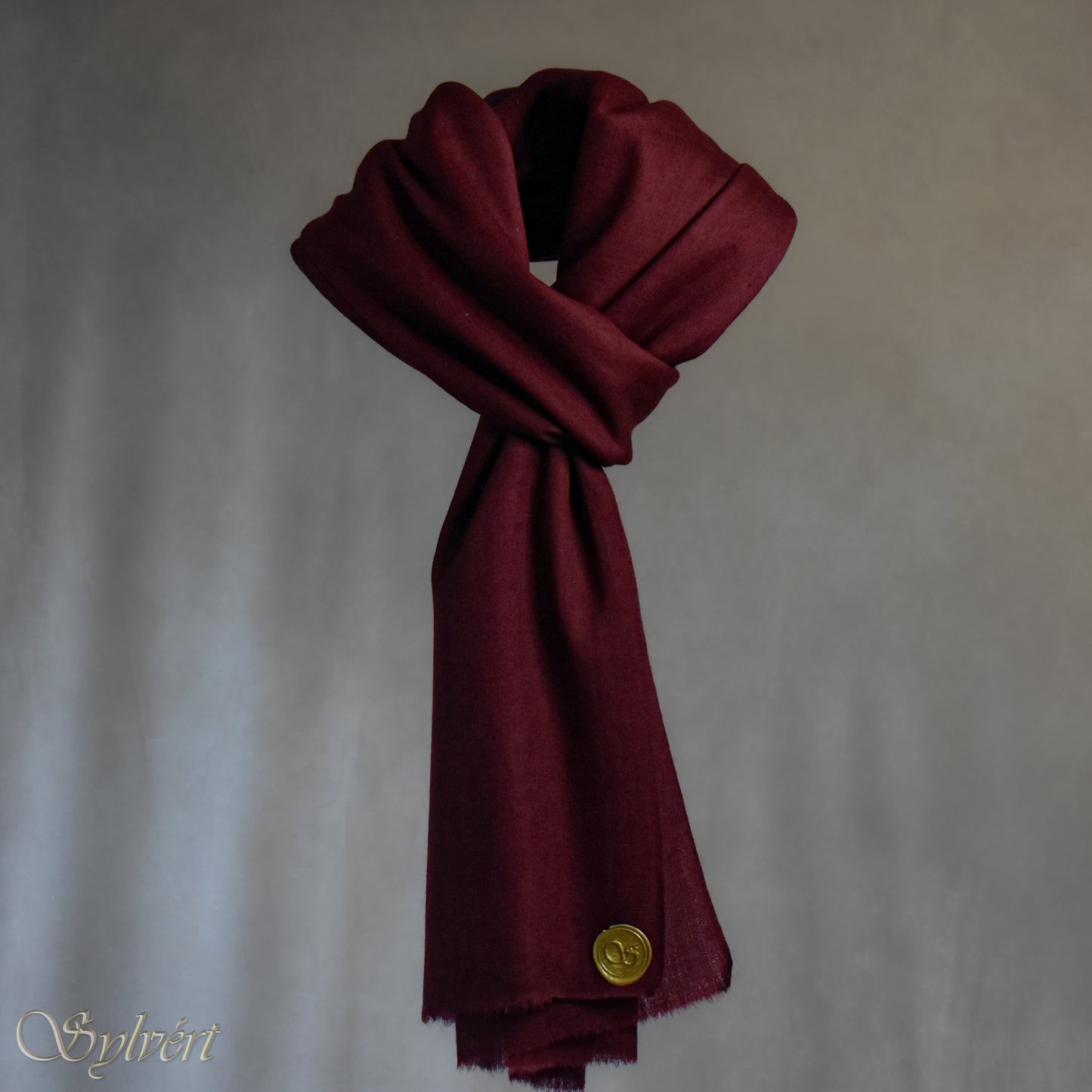 A maroon cashmere Rivella Morven Scarf from Sylvért's Rivanor Collection, looped and threaded showing the gold Sylvért stamp, suspended in the air in front to of a beige background