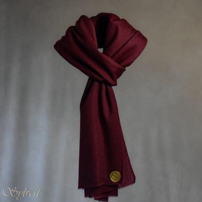 A maroon cashmere Rivella Morven Scarf from Sylvért's Rivanor Collection, looped and threaded showing the gold Sylvért stamp, suspended in the air in front to of a beige background