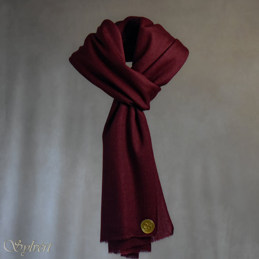 A maroon cashmere Rivella Morven Scarf from Sylvért's Rivanor Collection, looped and threaded showing the gold Sylvért stamp, suspended in the air in front to of a beige background
