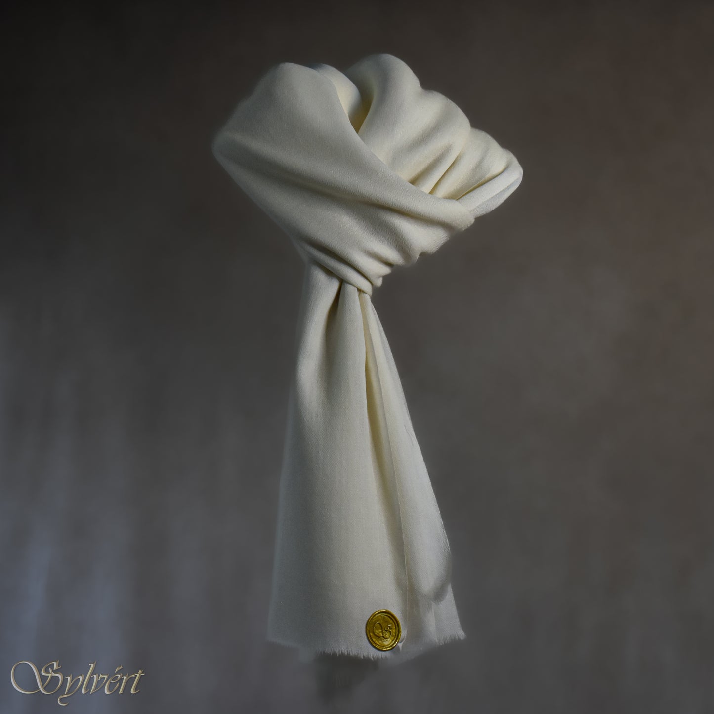 A white cashmere Rivella Caelis Scarf from Sylvért's Rivanor Collection, looped and threaded showing the gold Sylvért stamp, suspended in the air in front to of a beige background