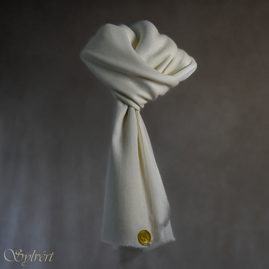 A white cashmere Rivella Caelis Scarf from Sylvért's Rivanor Collection, looped and threaded showing the gold Sylvért stamp, suspended in the air in front to of a beige background
