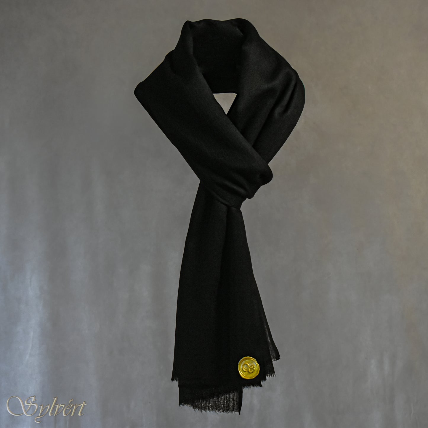 A black cashmere Rivella Velin Scarf from Sylvért's Rivanor Collection, looped and threaded showing the gold Sylvért stamp, suspended in the air in front to of a beige background