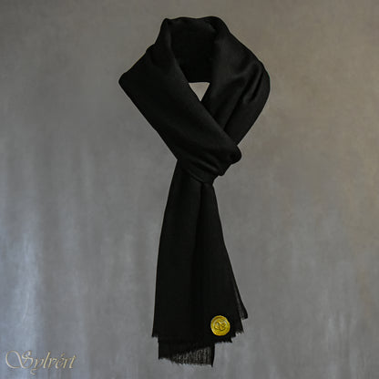 A black cashmere Rivella Velin Scarf from Sylvért's Rivanor Collection, looped and threaded showing the gold Sylvért stamp, suspended in the air in front to of a beige background