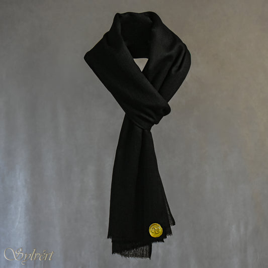 A black cashmere Rivella Velin Scarf from Sylvért's Rivanor Collection, looped and threaded showing the gold Sylvért stamp, suspended in the air in front to of a beige background