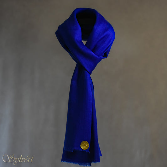 A blue cashmere Rivella Orven Scarf from Sylvért's Rivanor Collection, looped and threaded showing the gold Sylvért stamp, suspended in the air in front to of a beige background