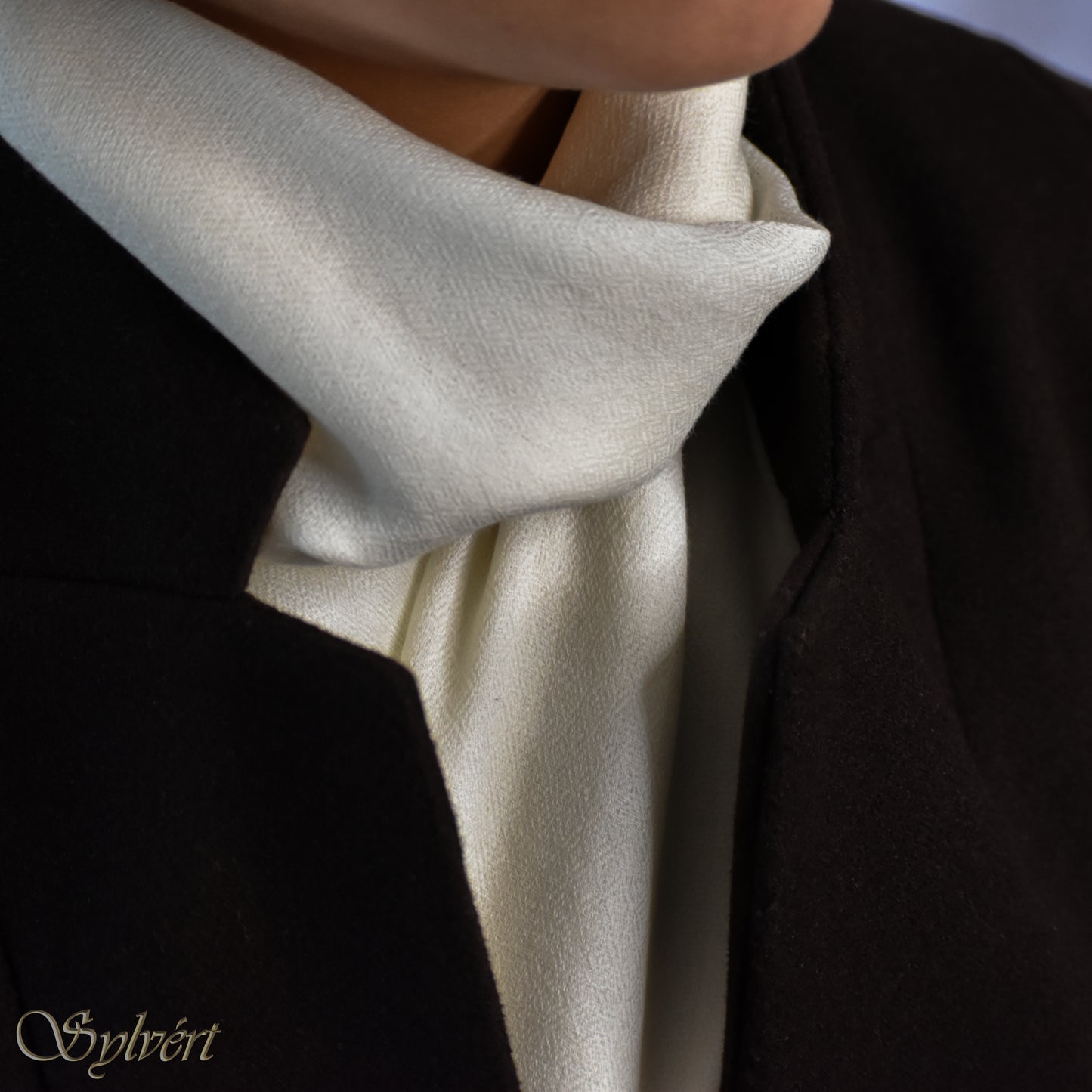 A styled shot of a white cashmere Rivella Caelis scarf from Sylvért's Rivanor collection worn under a dark brown coat.
