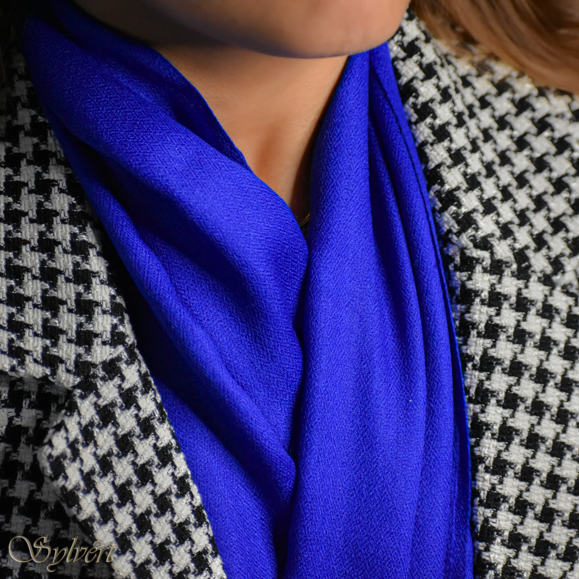 A styled shot of a blue cashmere Rivella Orven scarf from Sylvért's Rivanor collection worn under a black and white lattice jacket