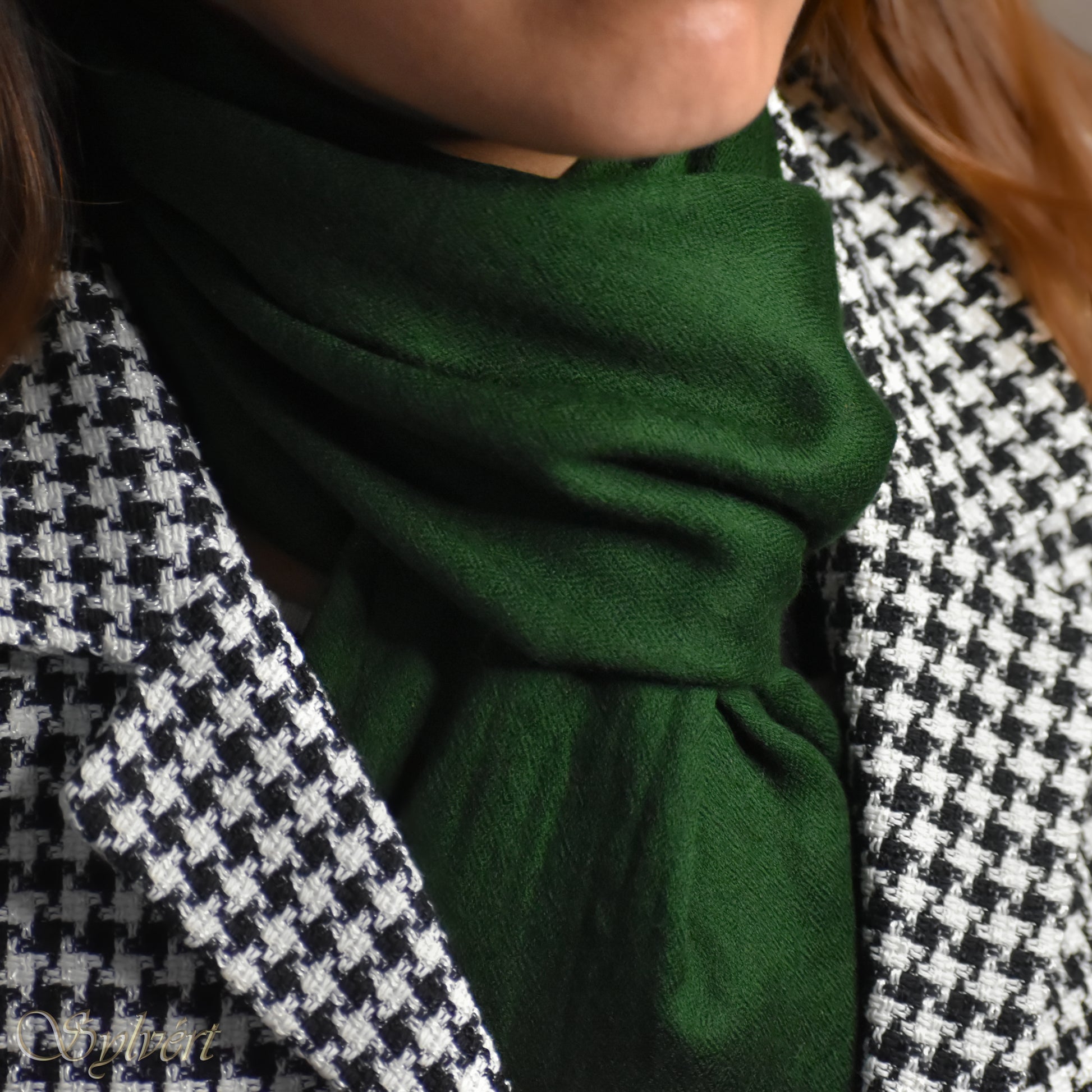 A styled shot of a green cashmere Rivella Tavon scarf from Sylvért's Rivanor collection worn under a black and white lattice jacket