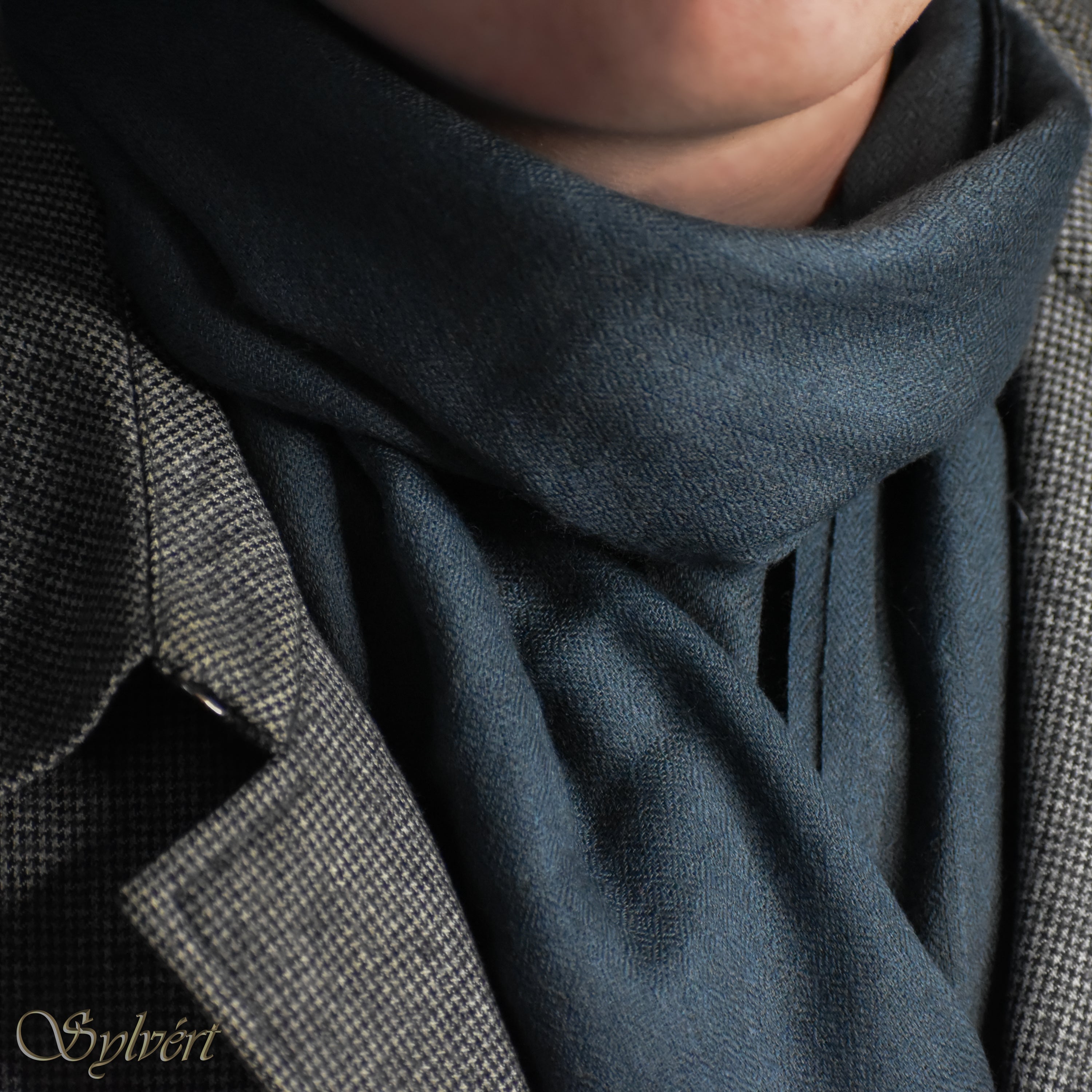 A styled shot of a blue-grey slate cashmere Rivella Morven scarf from Sylvért's Rivanor collection worn under a grey and black lattice coat